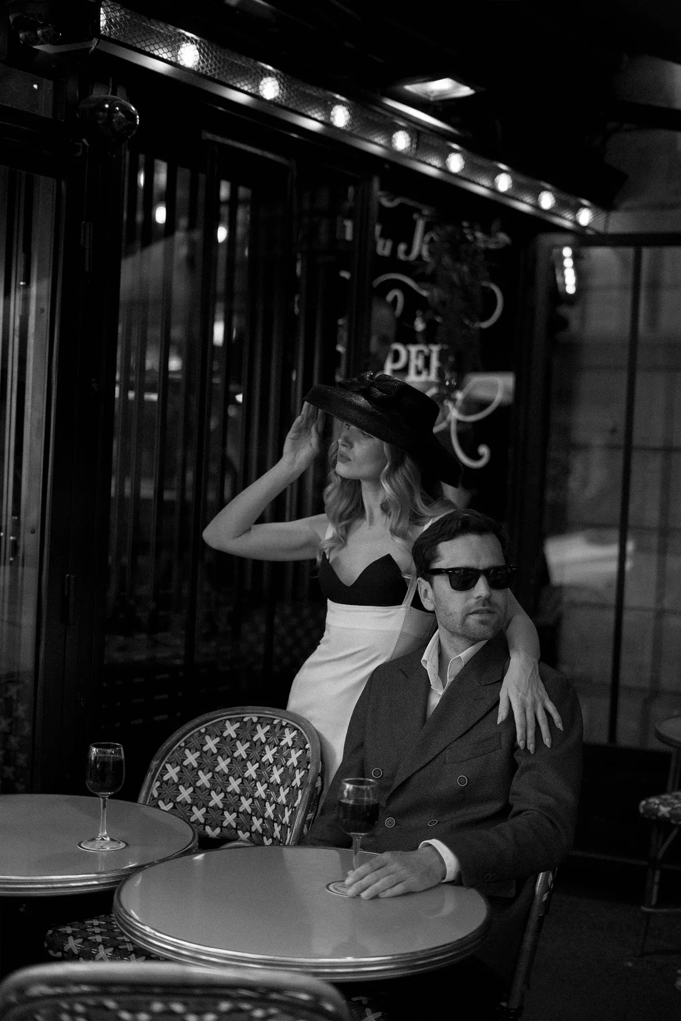 Couple at a sidewalk cafe at night, she adjusts a wide black hat behind his chair