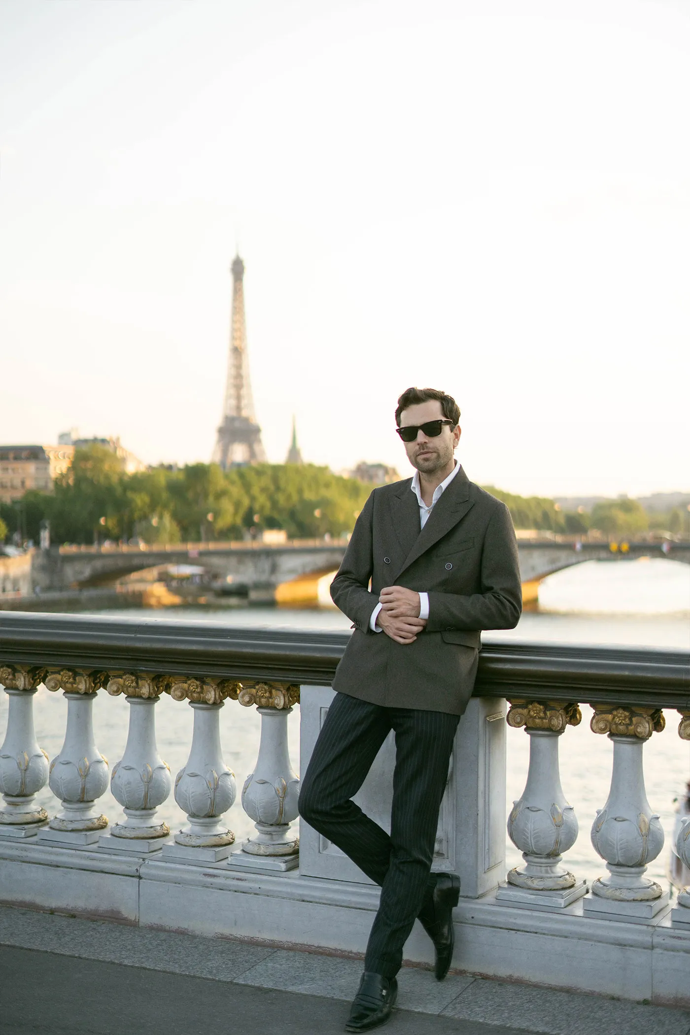 Man in a suit and sunglasses leaning on a bridge railing, Eiffel Tower behind