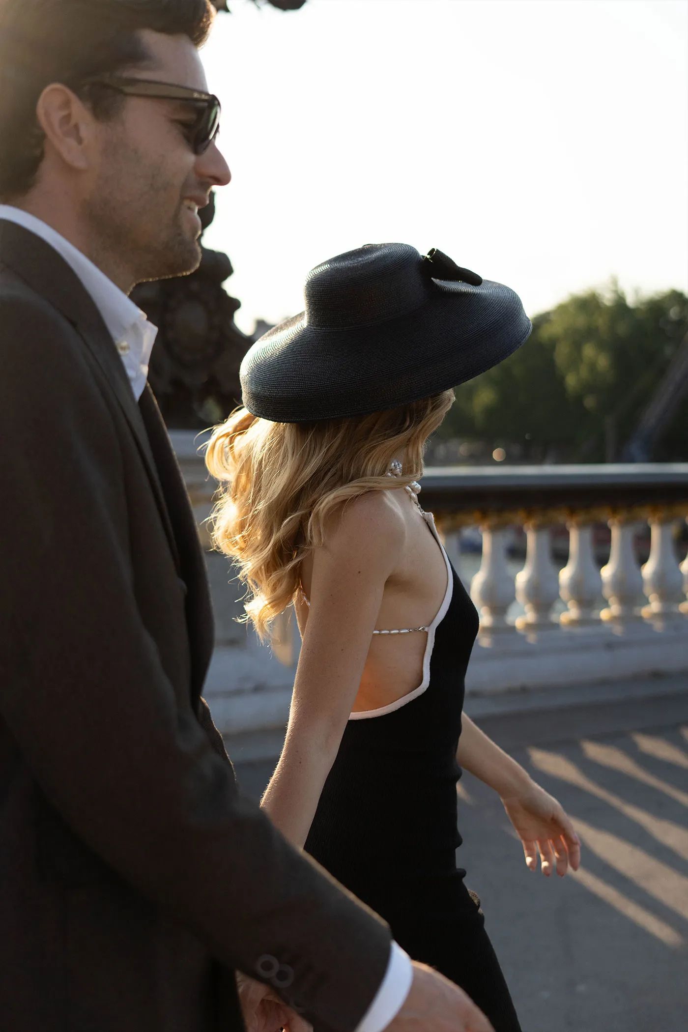 Woman in a black dress and wide-brimmed hat walking a bridge beside a man in a suit