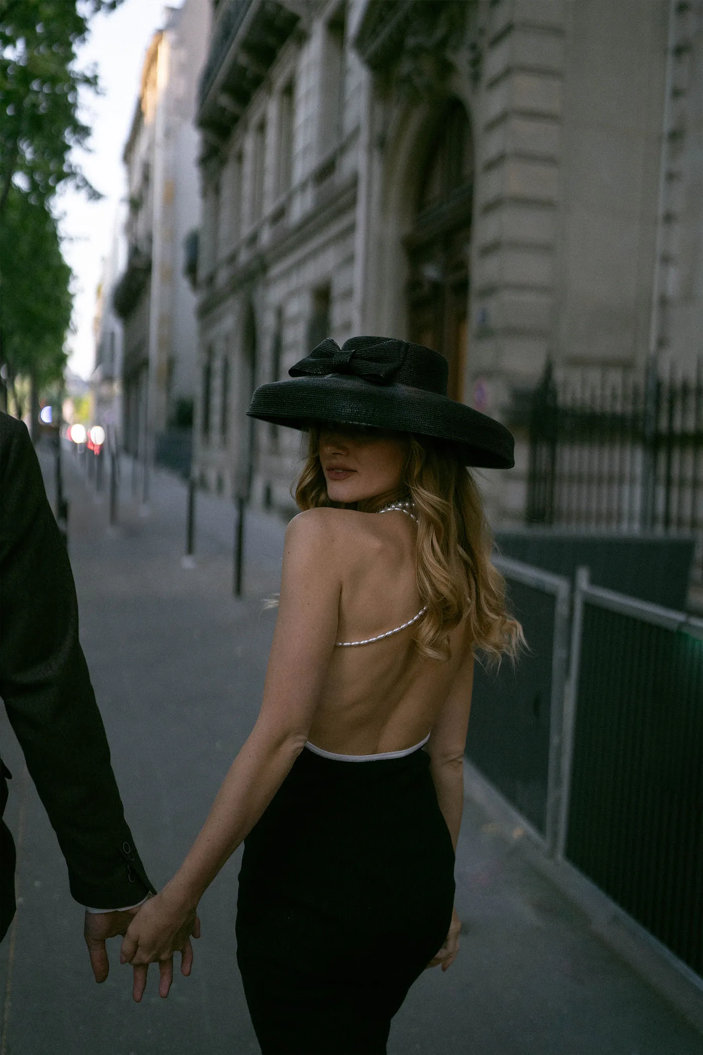 Woman in a black hat and backless dress holding hands on a city sidewalk