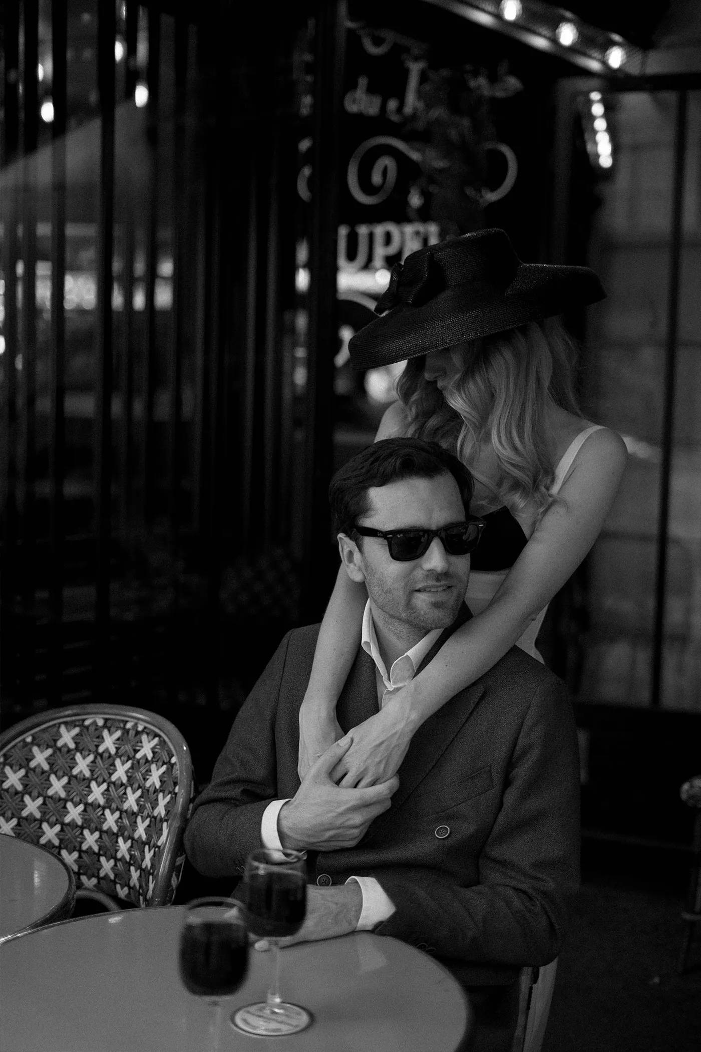 Woman in a black hat with her arms around a seated man at a cafe, wine glasses on the table