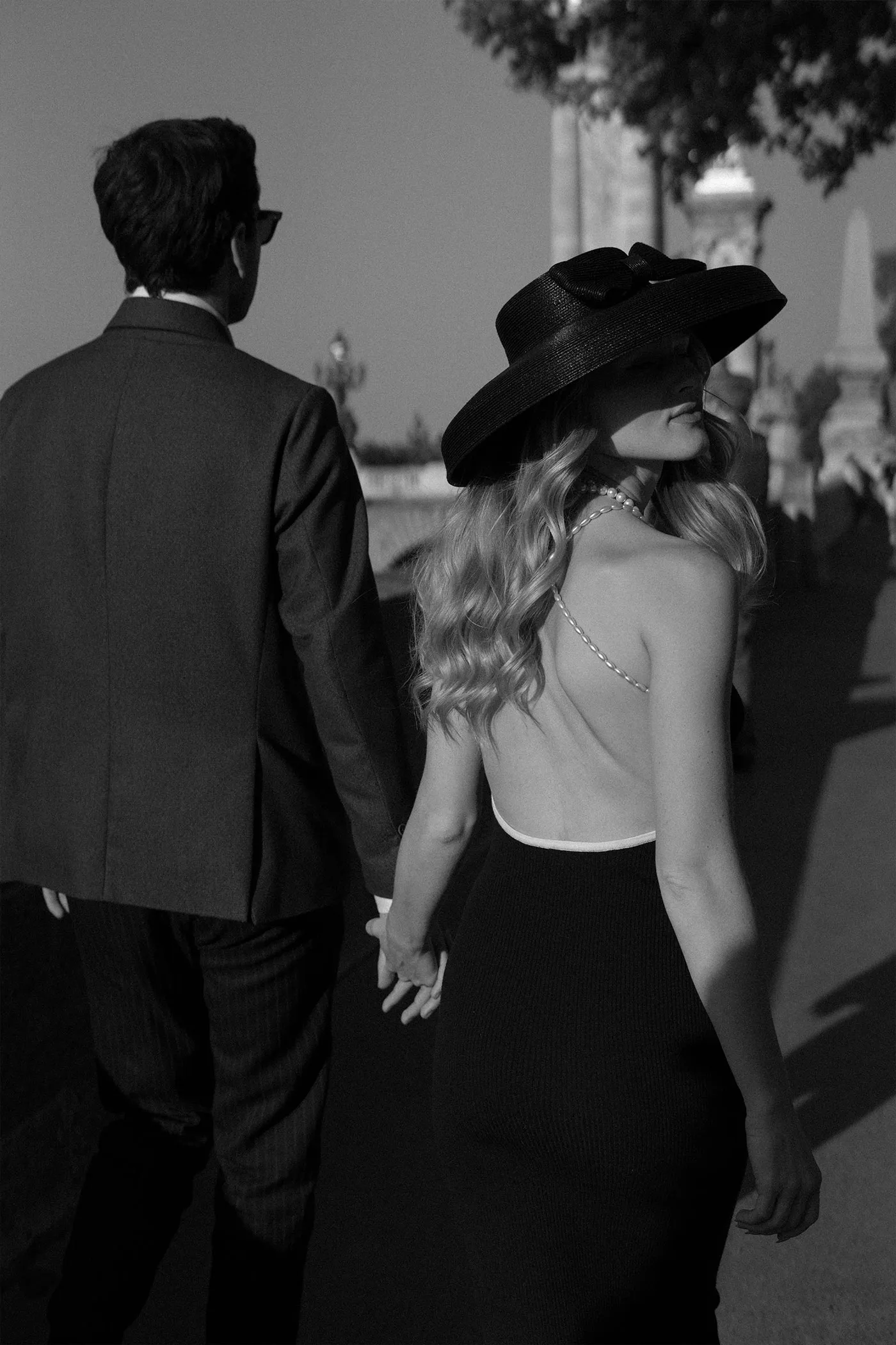 Woman in a wide black hat glancing back while holding a man's hand on a sunlit bridge