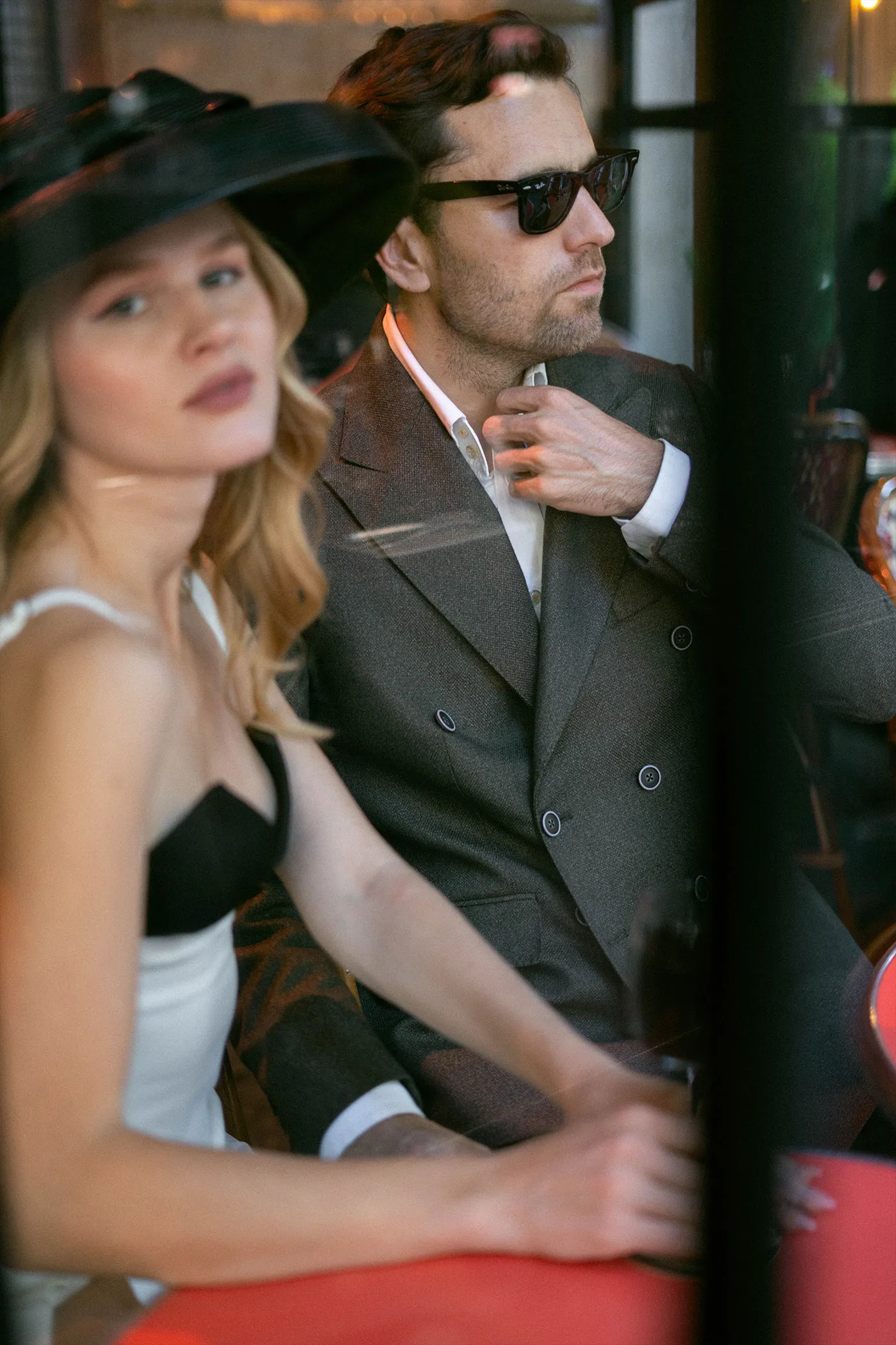 Woman in a wide-brimmed black hat and man in a tweed suit and sunglasses, seen through glass