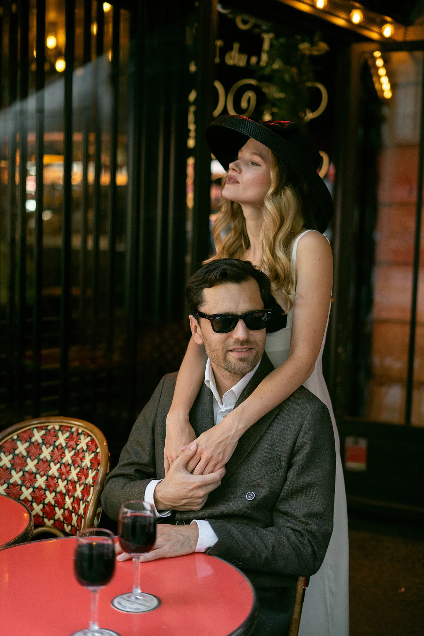 Woman in a wide black hat standing behind a man in sunglasses at a red cafe table