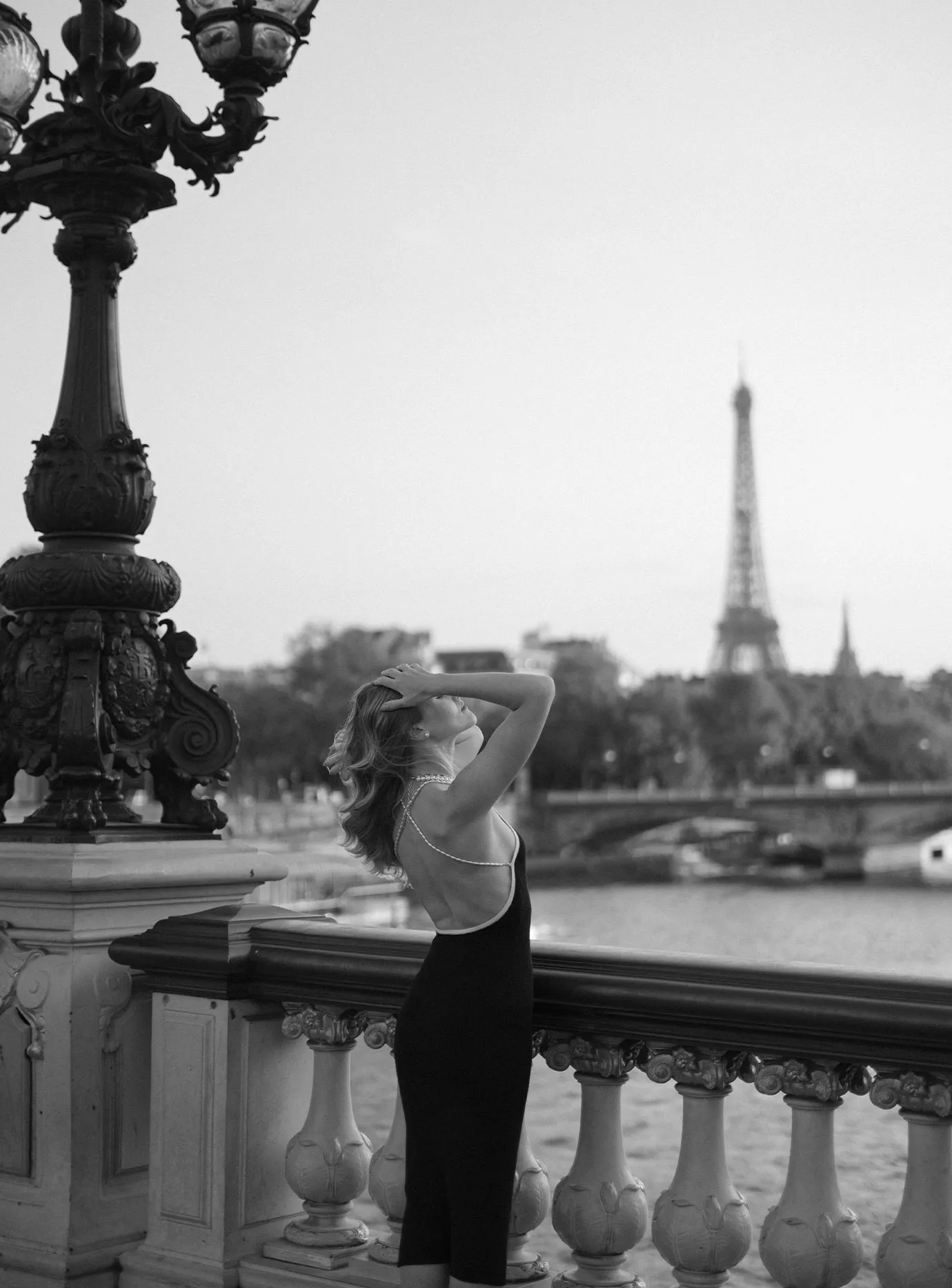 Woman in a black slip dress leaning on a bridge railing, Eiffel Tower behind her