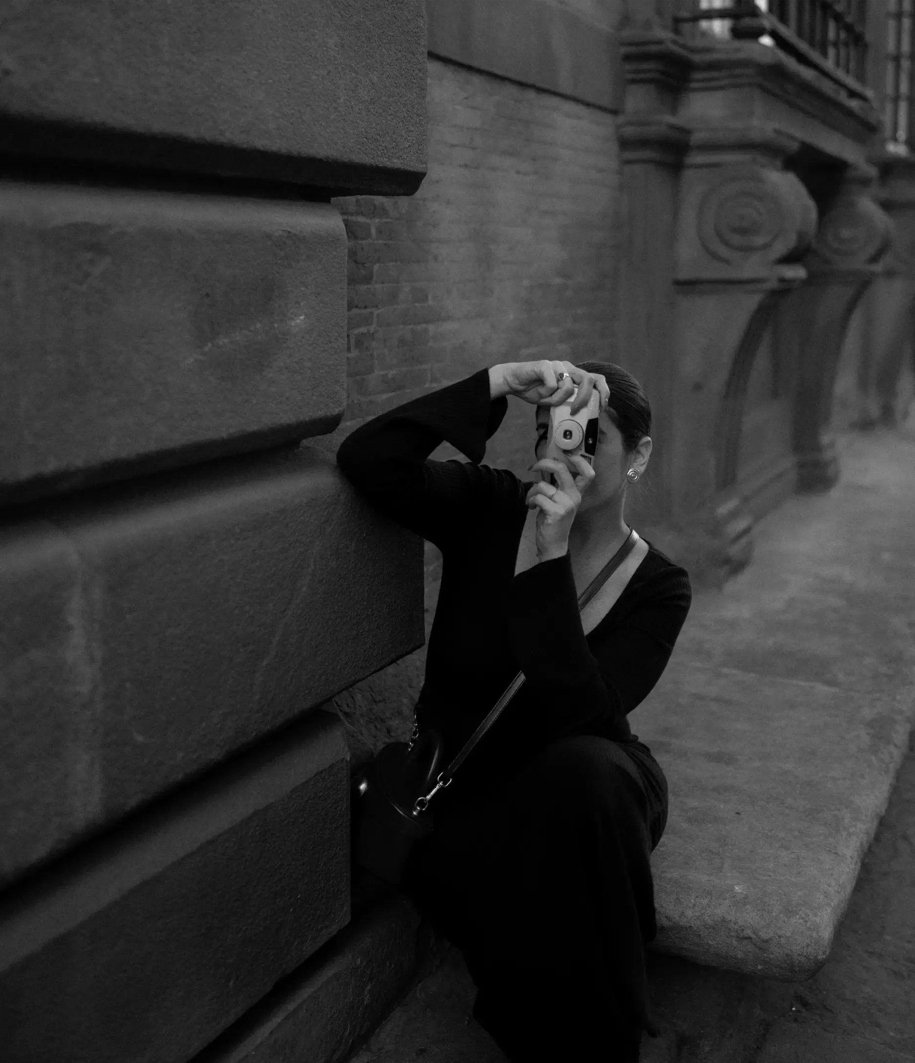 Photographer in black seated on a stone ledge, raising a film camera to her eye