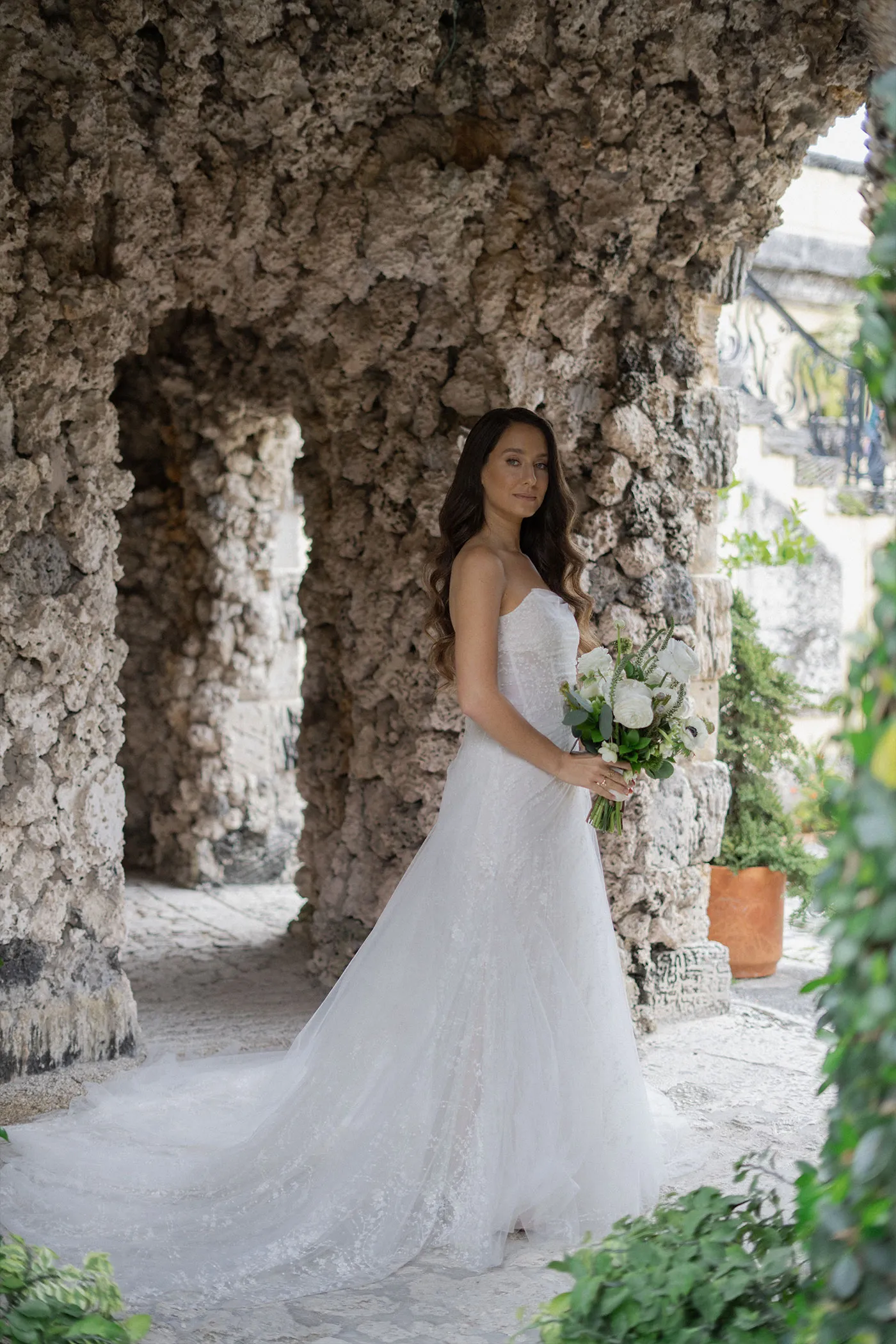 Bride holding a white bouquet under coral-stone arches, train spread behind her