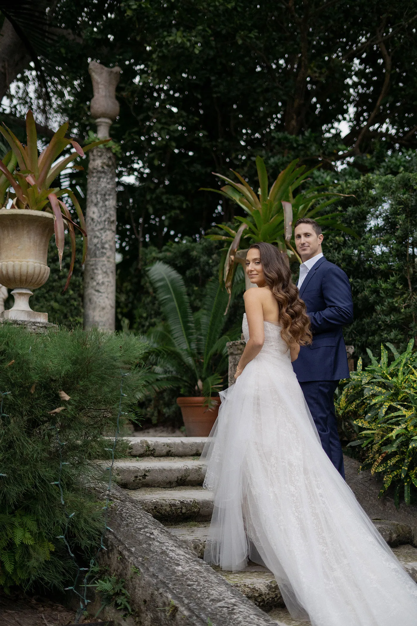 Bride glancing back over her shoulder on garden steps, groom in navy suit behind her