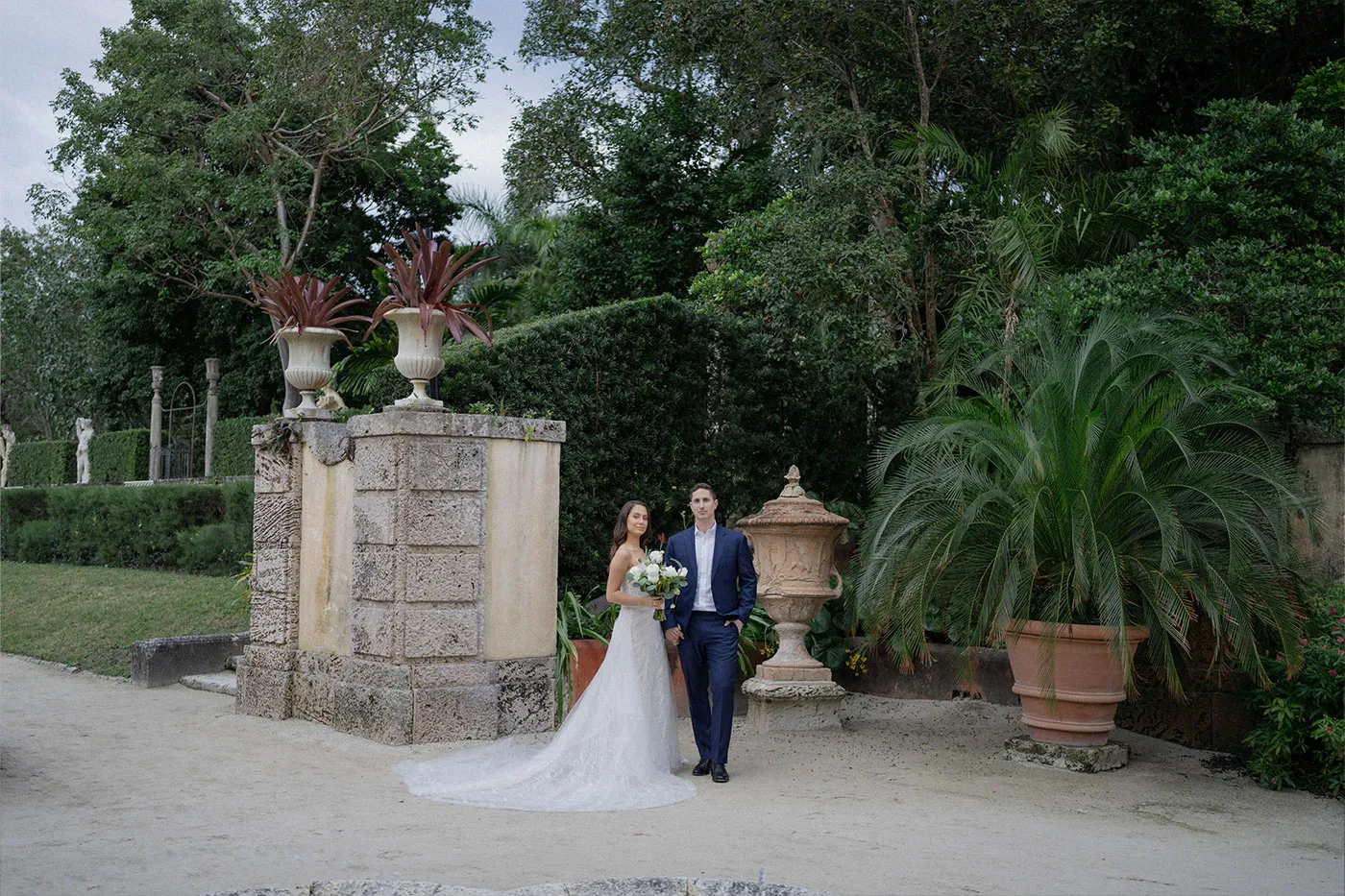 Bride with a bouquet and groom in navy standing by a stone pillar in a tropical garden