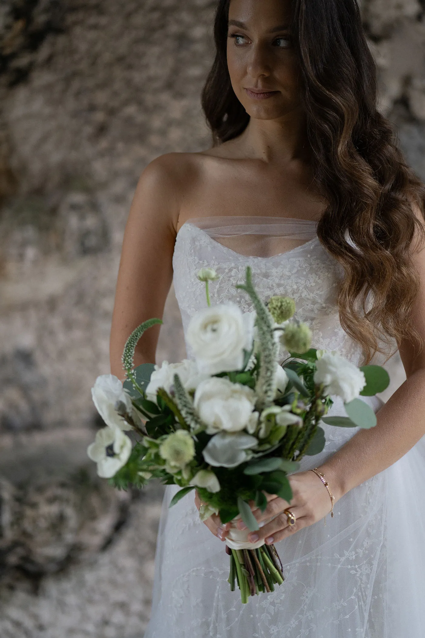 Bride in a lace gown holding a white ranunculus and anemone bouquet by a stone wall