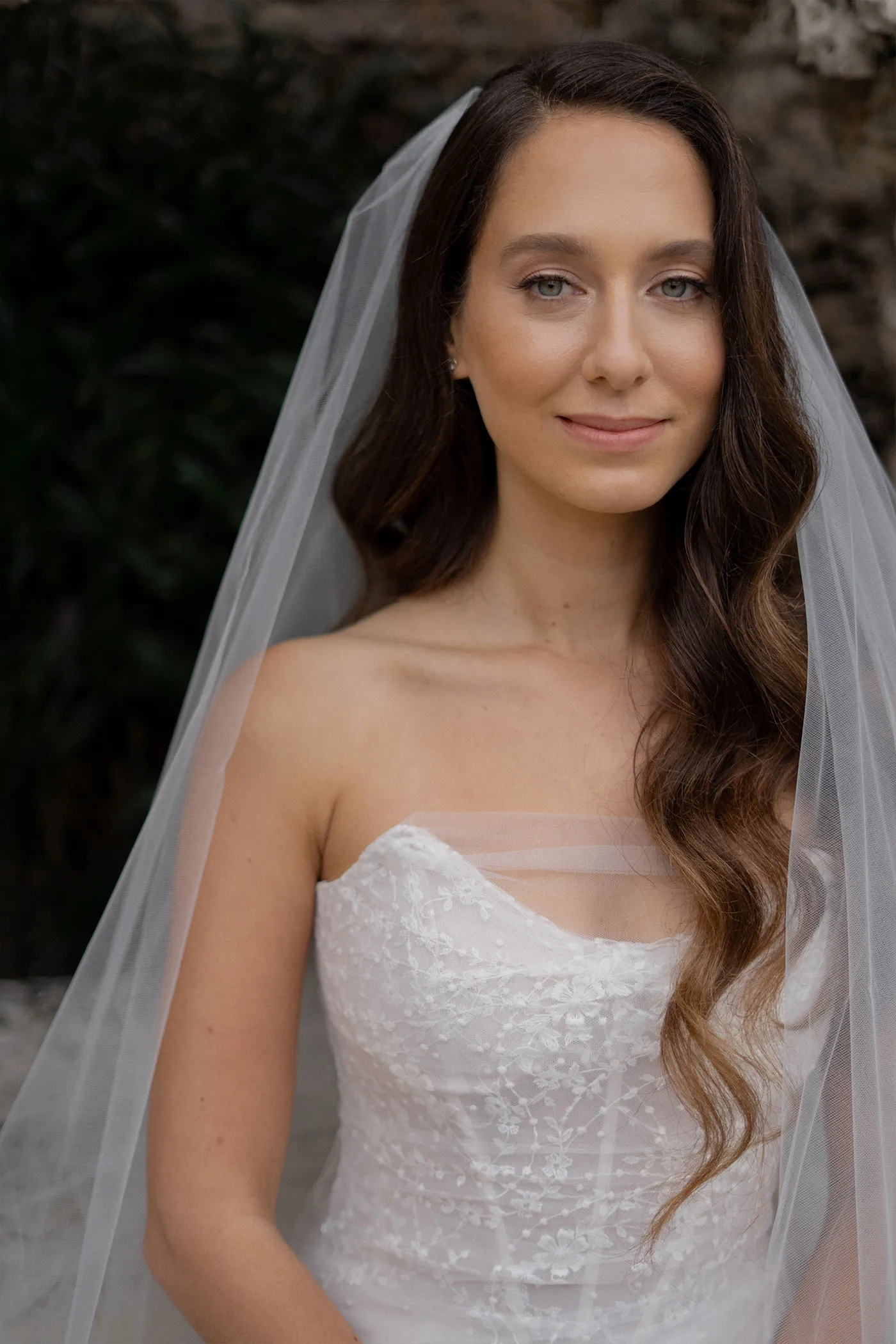 Bride in a lace gown and long veil, half smiling against dark foliage