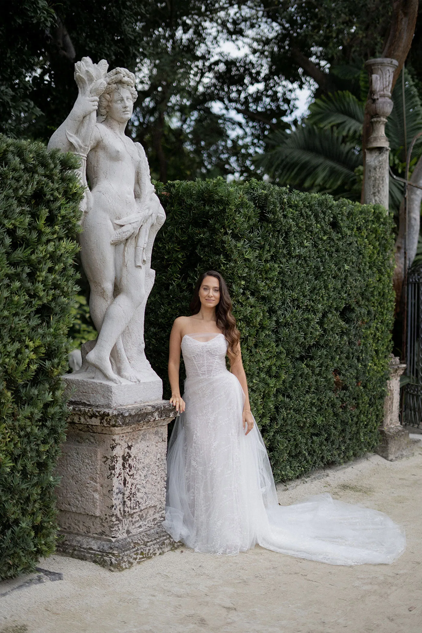 Bride in a strapless lace gown beside a stone statue and clipped hedge in a garden