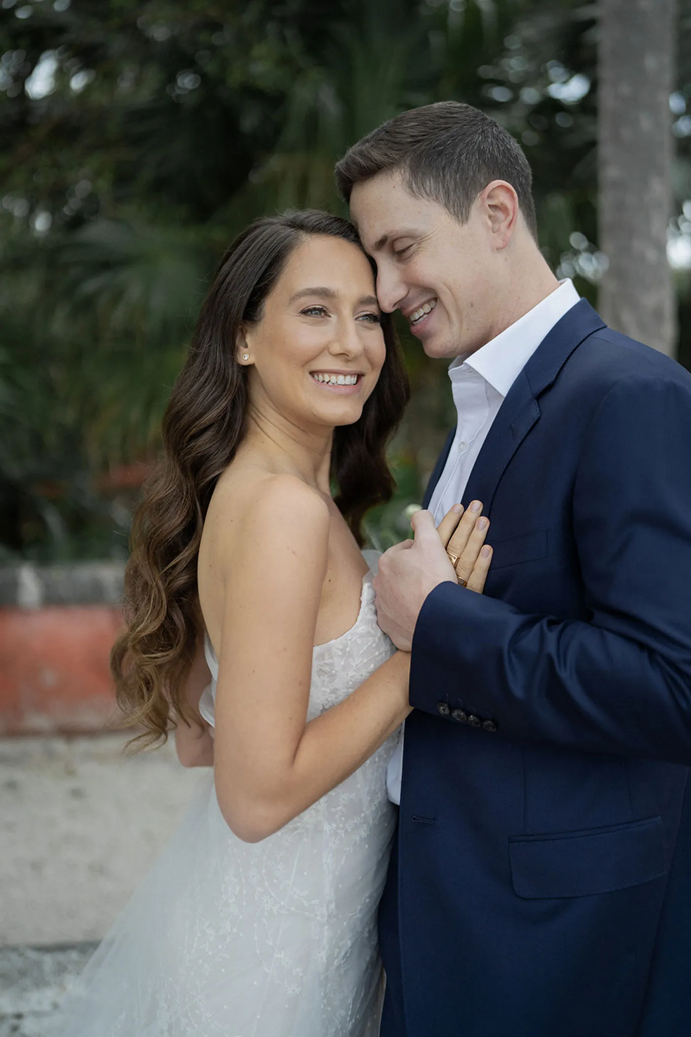 Bride in a strapless gown leaning into a groom in a navy suit, both laughing outdoors
