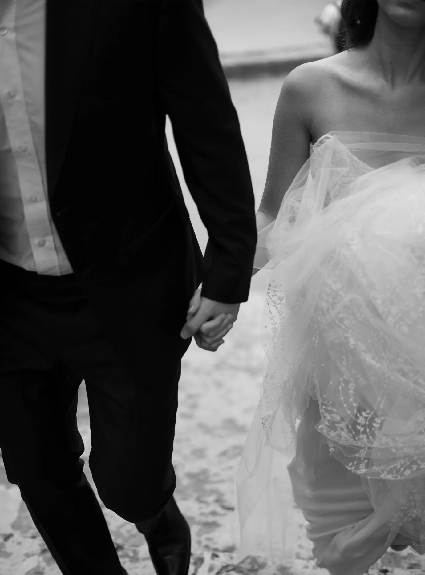 Bride in tulle and groom in a tuxedo holding hands as they walk across sand