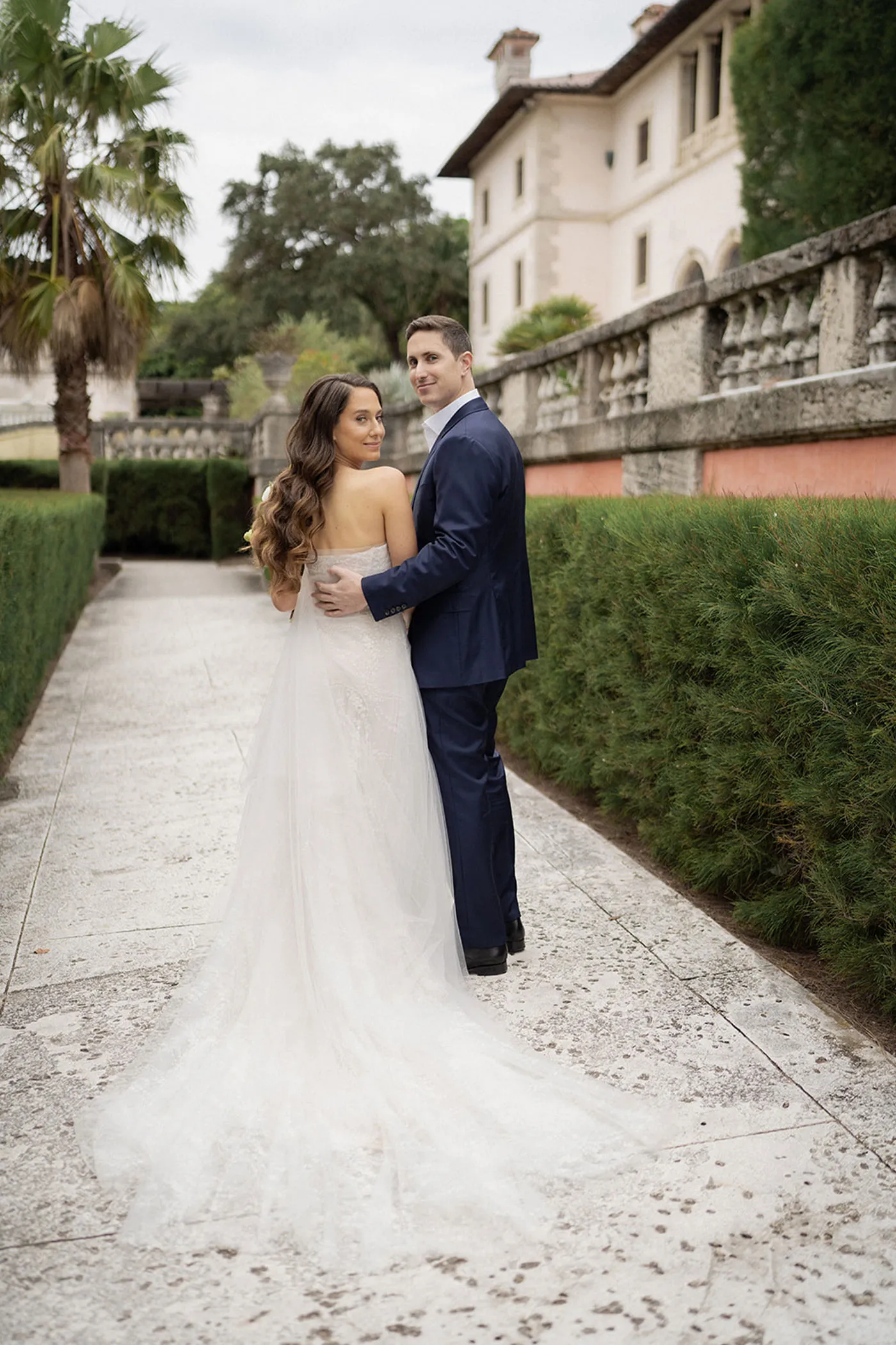 Bride with a long tulle train and groom in navy looking back on a hedge-lined garden path