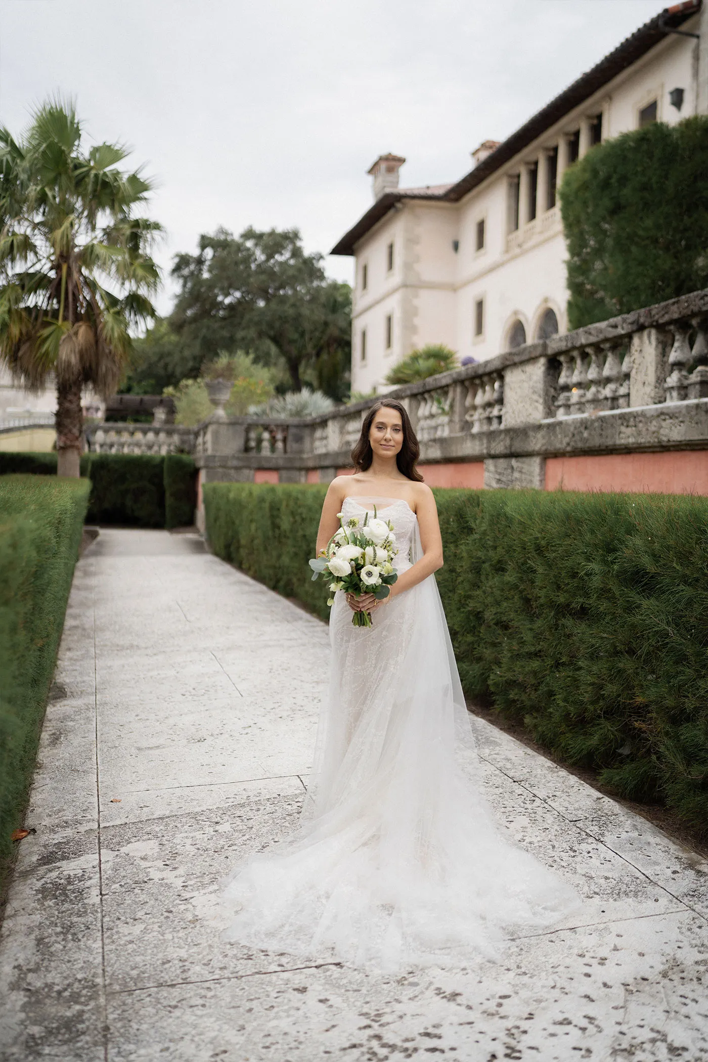 Bride holding white anemones on a hedge-lined path in front of a stone villa