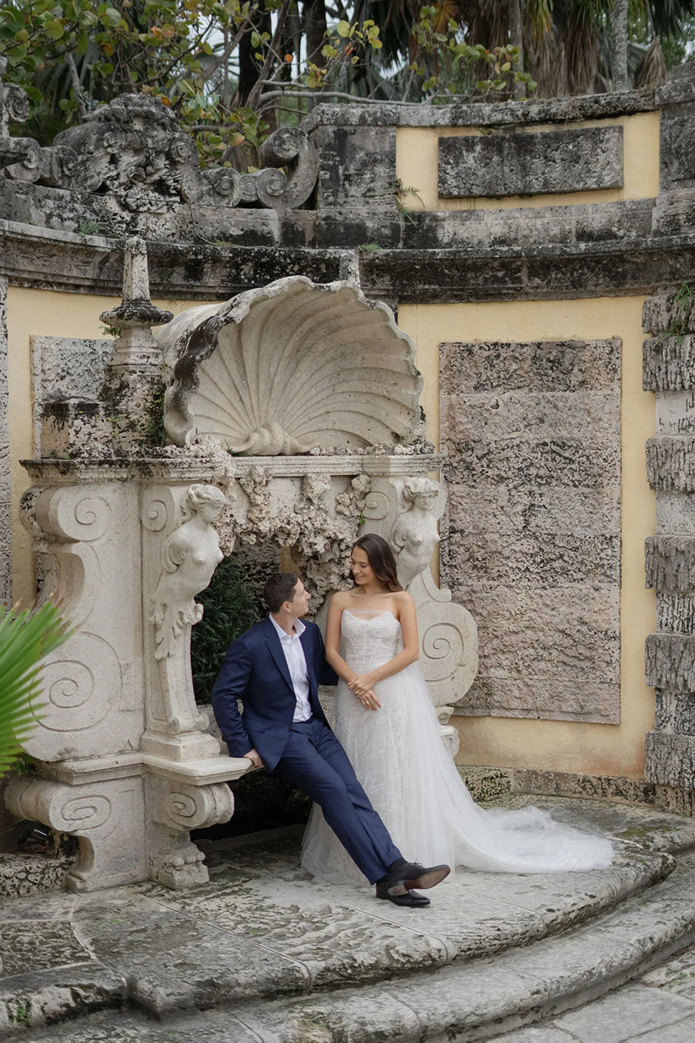 Bride and groom by a carved stone shell fountain in a weathered garden grotto