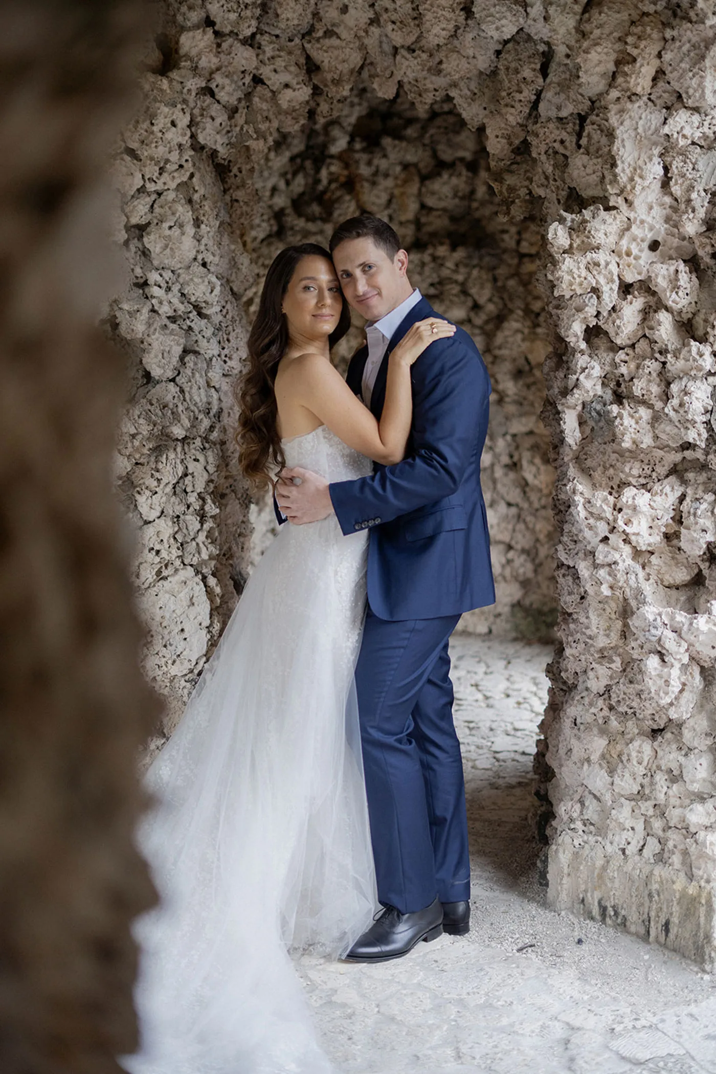 Bride and groom embracing in a coral-stone archway, groom in a navy suit