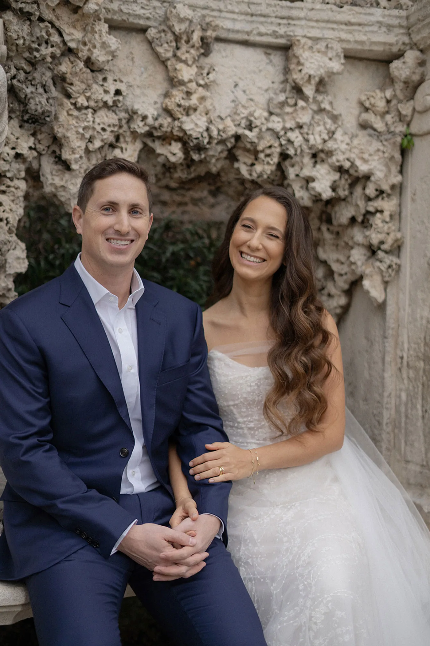 Bride and groom laughing side by side on a coral stone bench in a garden