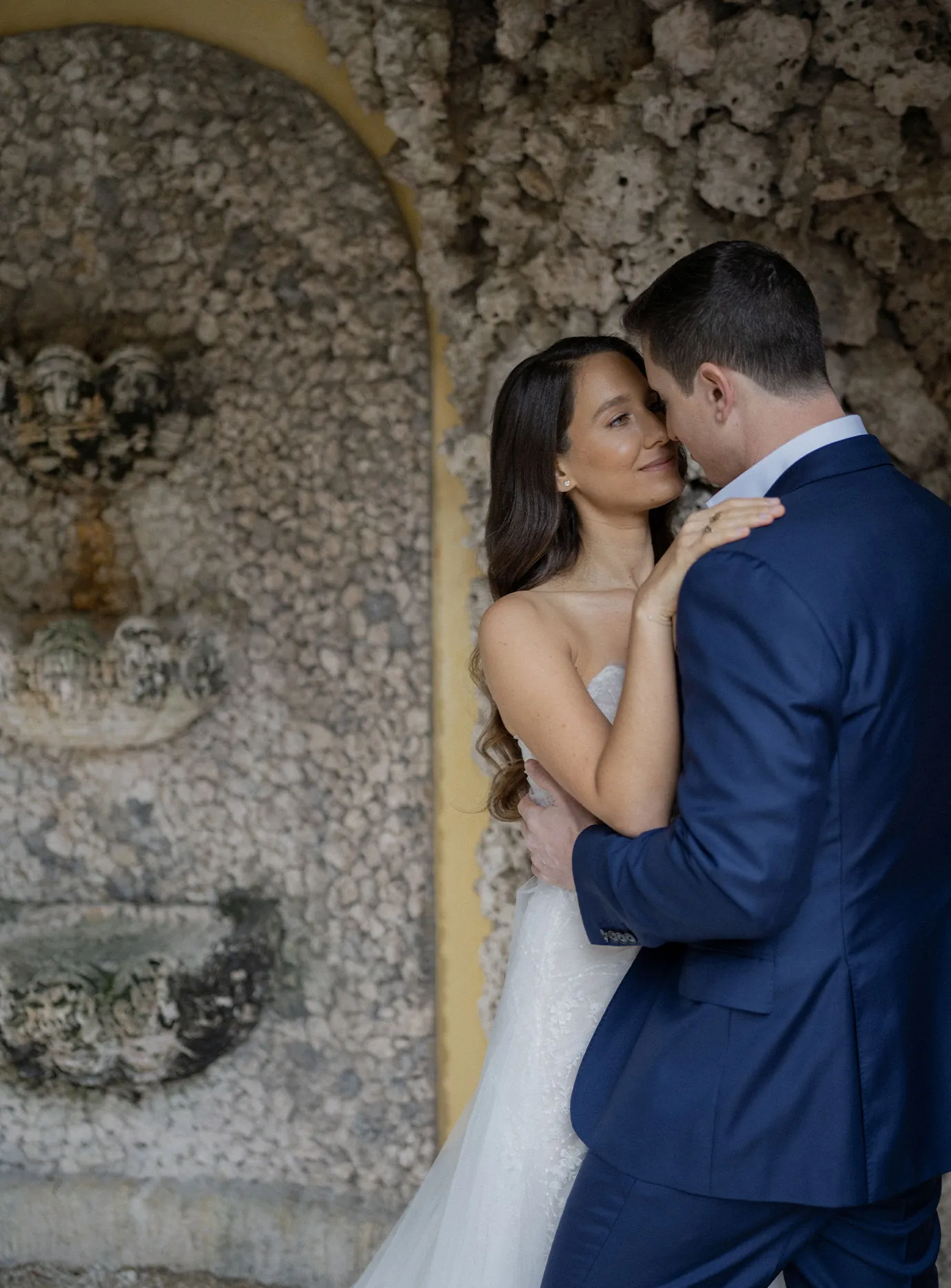 Bride and groom in a navy suit holding each other beside a coral rock grotto fountain