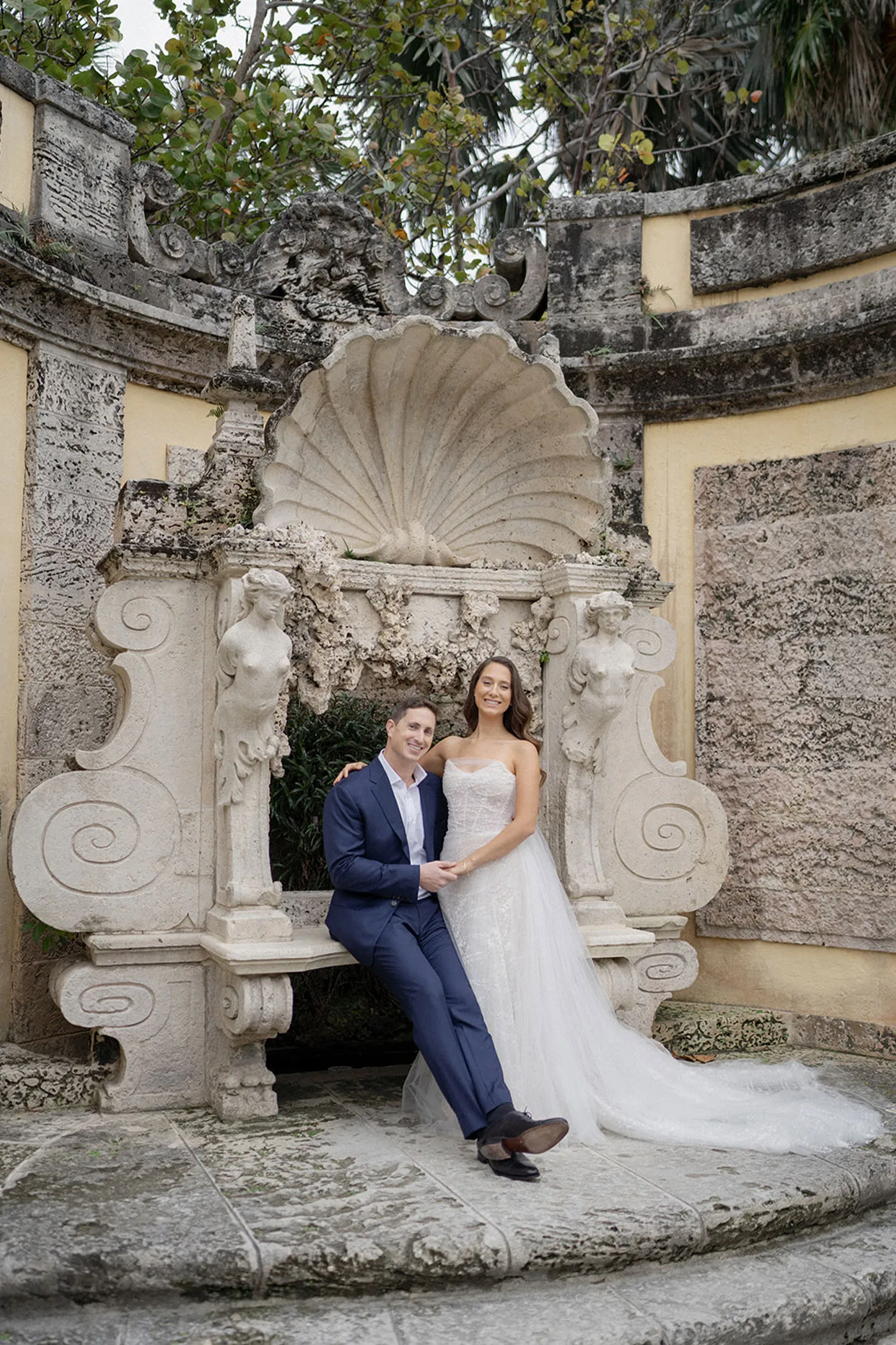 Bride and groom holding hands on a stone bench beneath a carved shell fountain