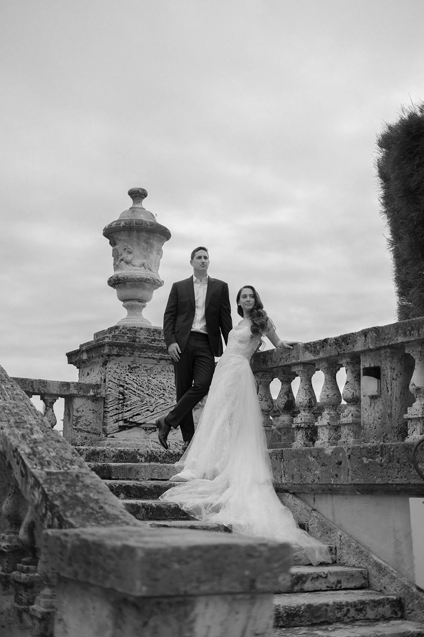 Bride and groom on weathered stone garden steps under an overcast sky, in black and white