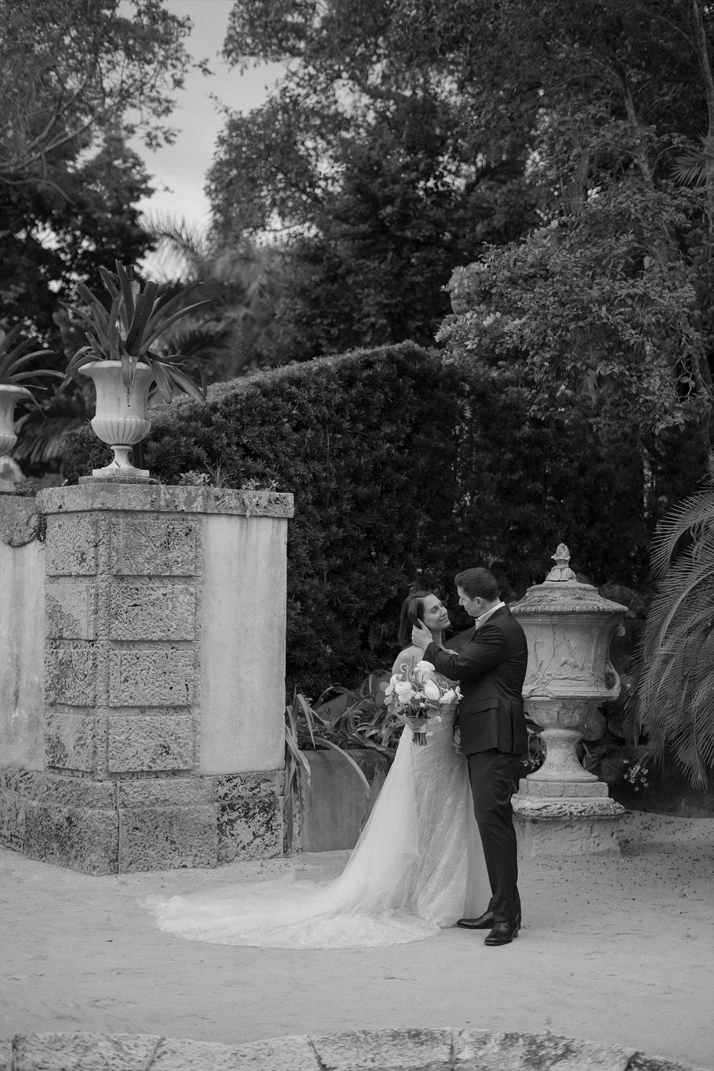 Bride and groom holding each other on a garden terrace between stone urns, black and white