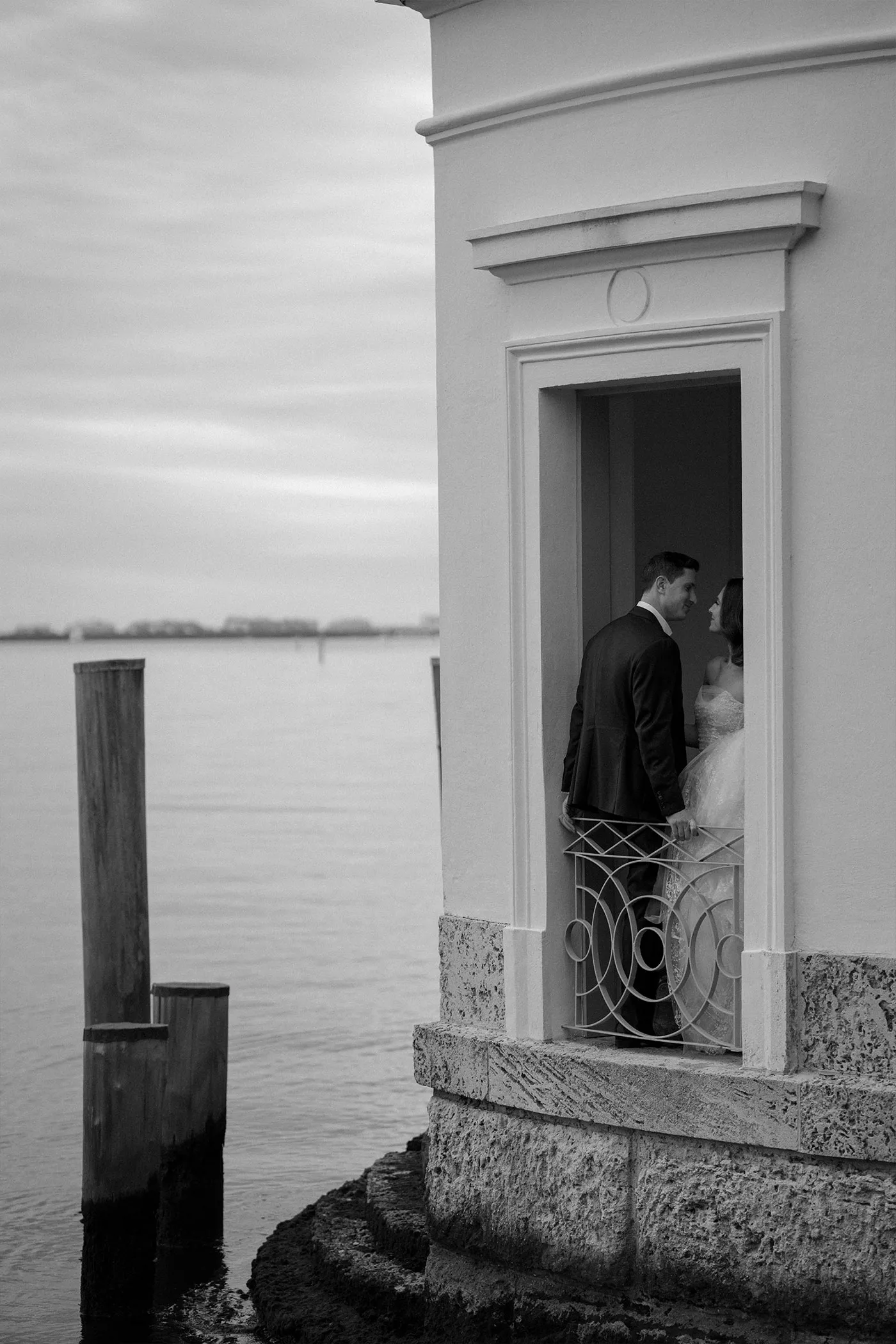 Bride and groom facing each other in a waterfront doorway above still bay water
