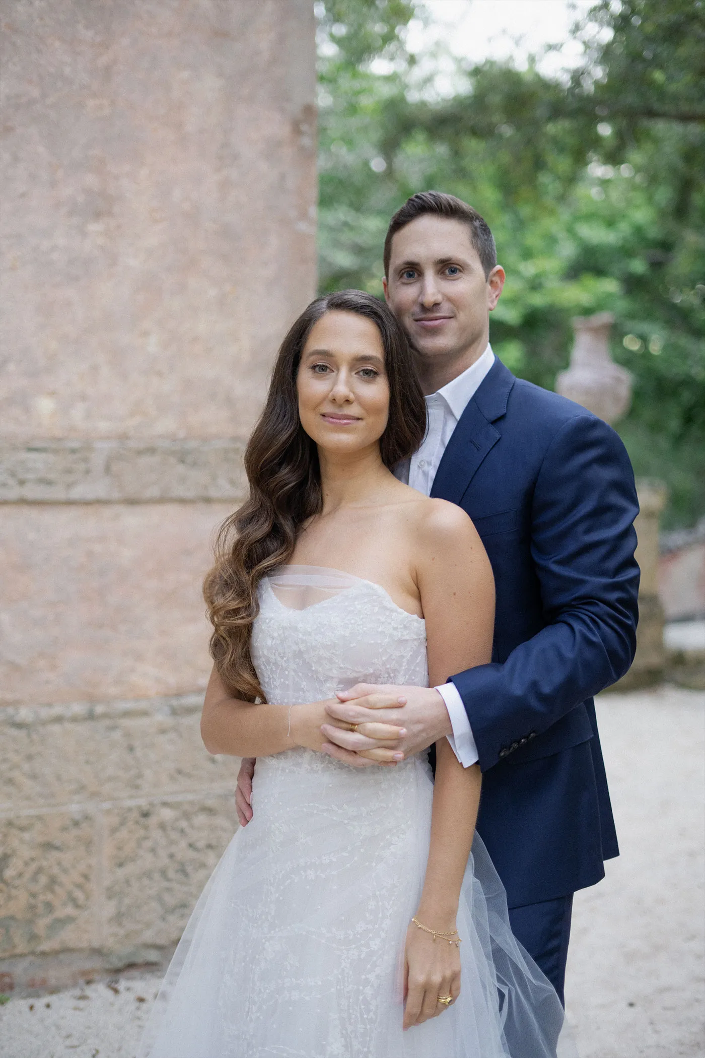 Groom in a navy suit holding the bride from behind against a weathered stone wall