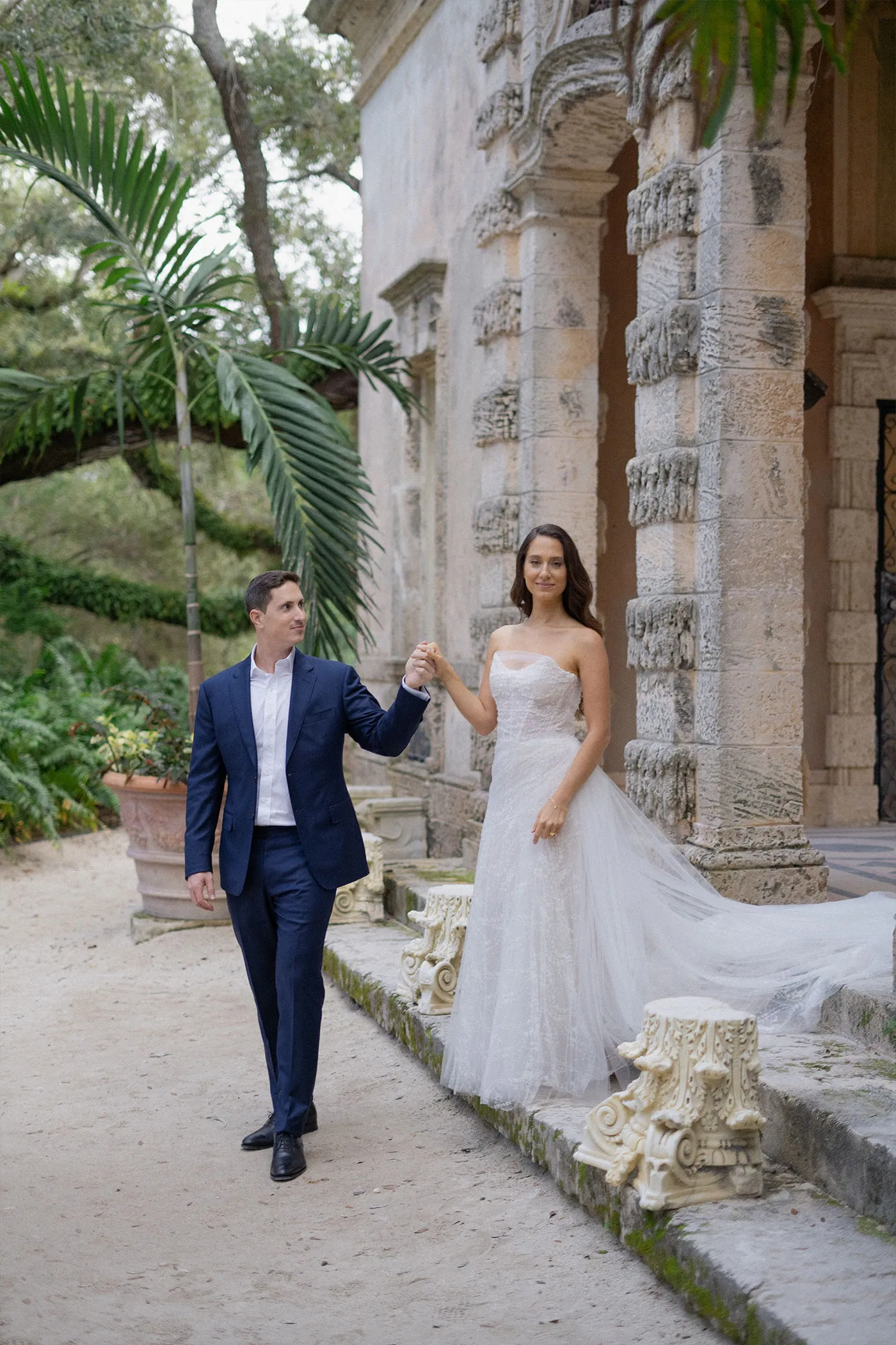 Groom leading the bride by the hand along a gravel path beside a stone villa