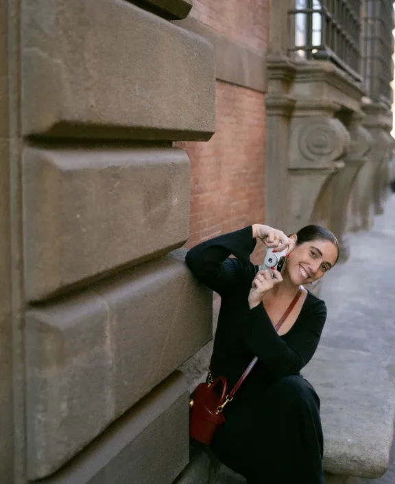 Photographer in black leaning on a stone building, raising a film camera and smiling