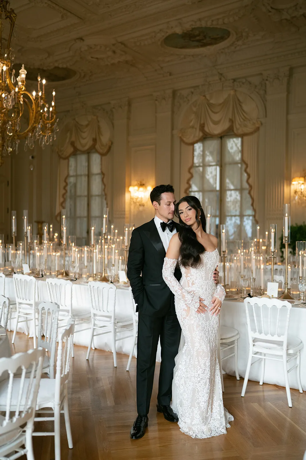 Bride in a beaded lace gown leaning on her groom in a candlelit ballroom