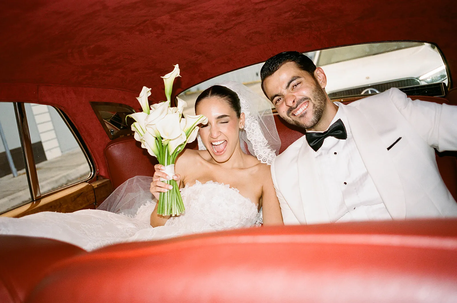 Bride with calla lilies laughing beside her groom in the back of a vintage car