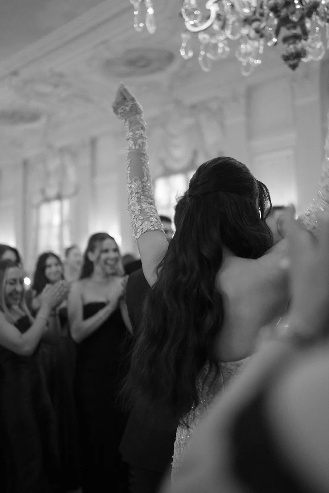 Bride with a raised arm dancing as guests clap around her, in black and white