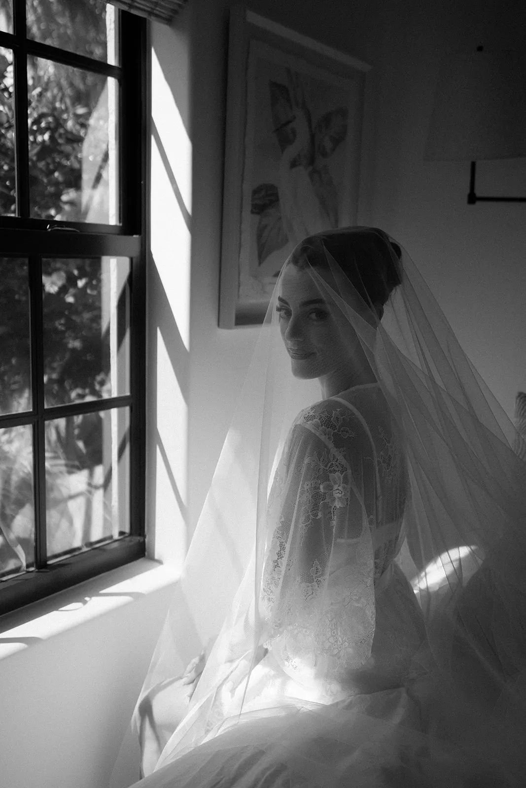 Bride under a long veil by a sunlit window, in black and white