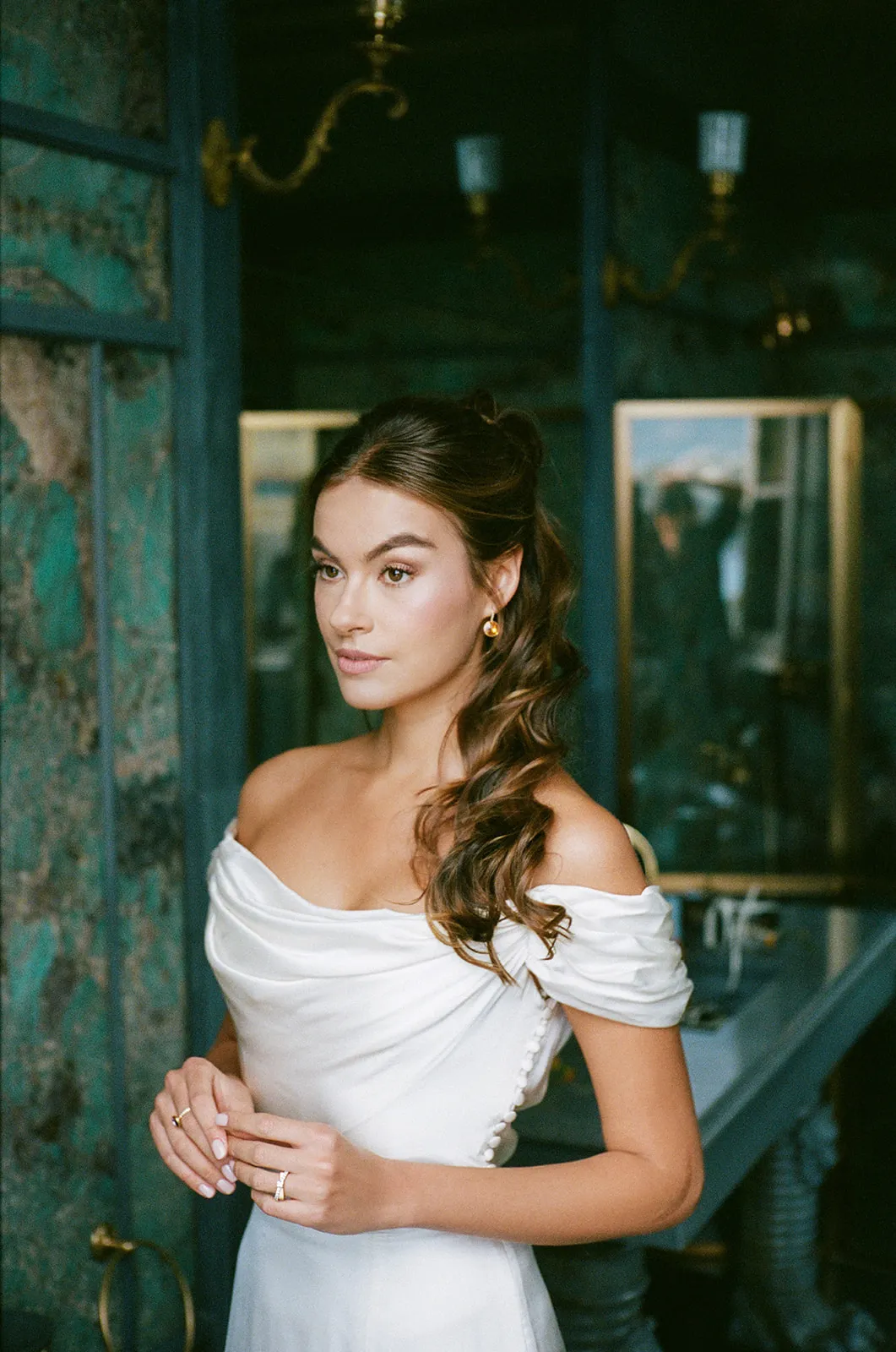 Bride in an off-shoulder gown standing by a teal-panelled wall with gilt mirrors