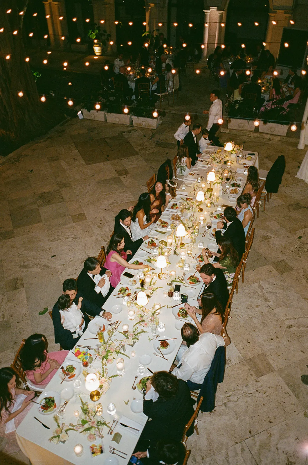 Long candlelit banquet table of guests in a stone courtyard under string lights