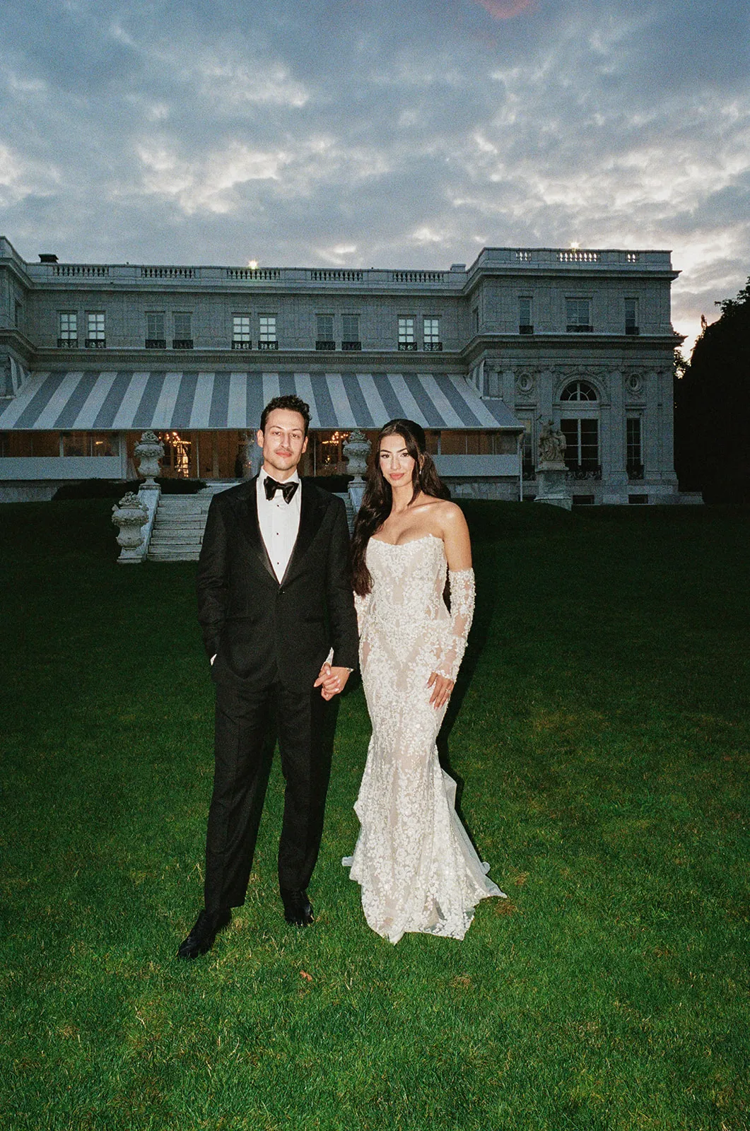 Bride and groom holding hands on a lawn before a grand mansion at dusk