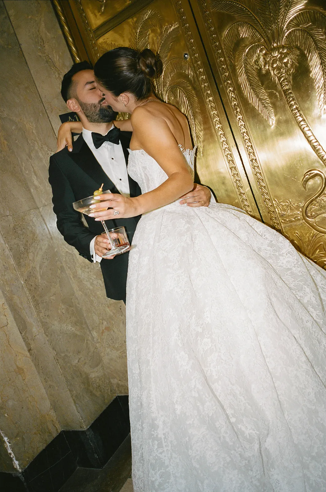 Couple kissing on marble stairs beside gilded doors, drinks in hand