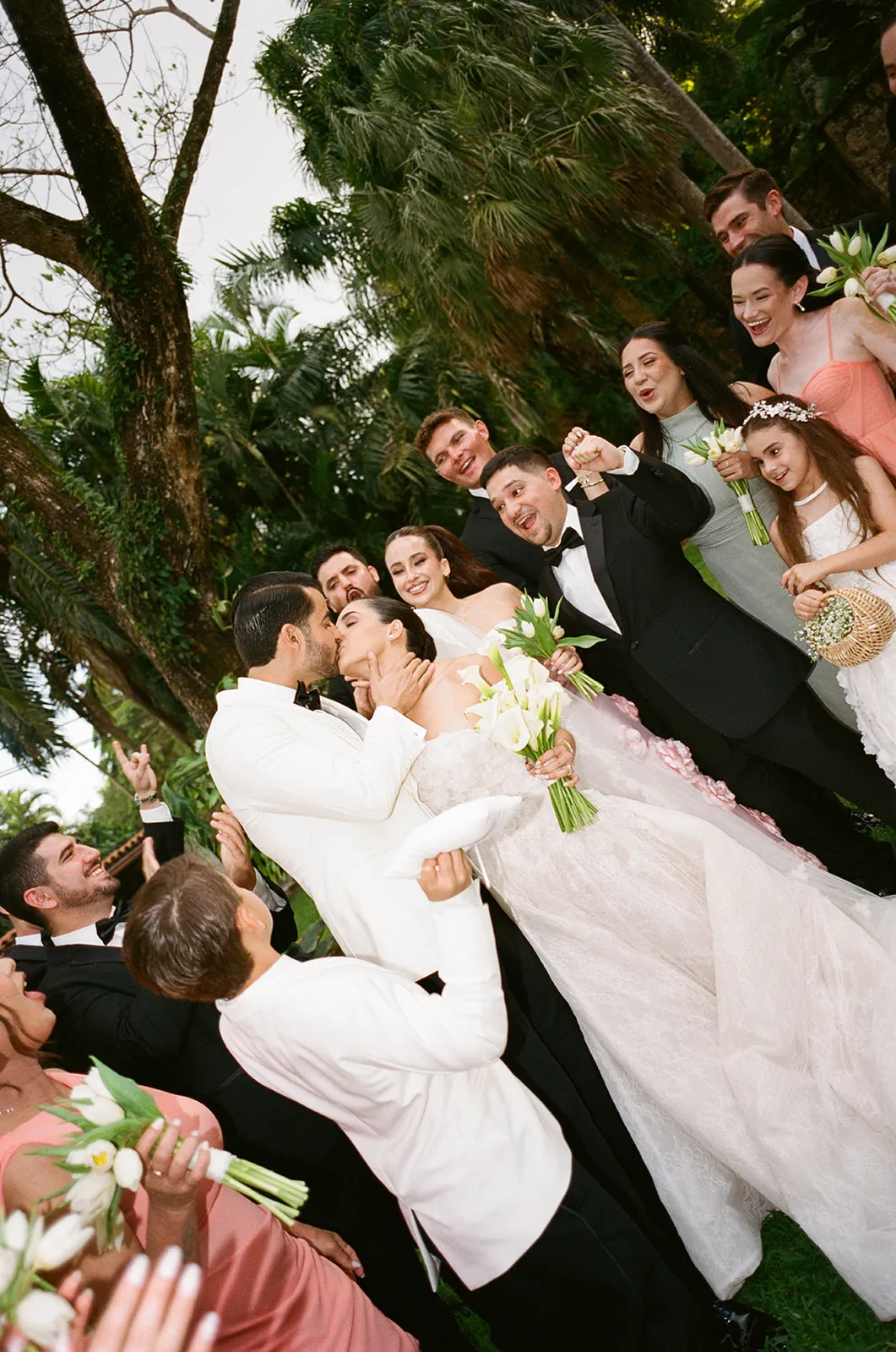 Couple kissing as the wedding party cheers around them under palm trees