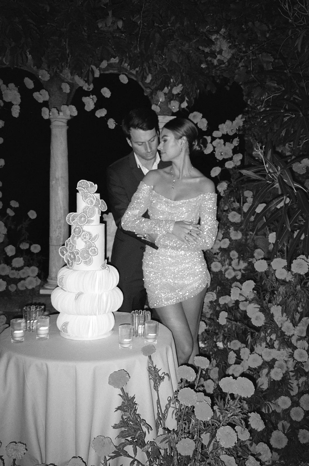 Couple beside a tiered wedding cake at night, the bride in a sequinned mini dress