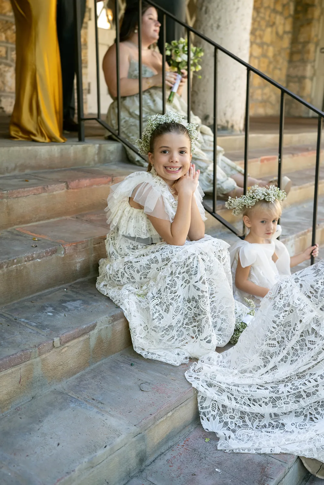 Two flower girls in lace dresses and baby's breath crowns sitting on stone steps