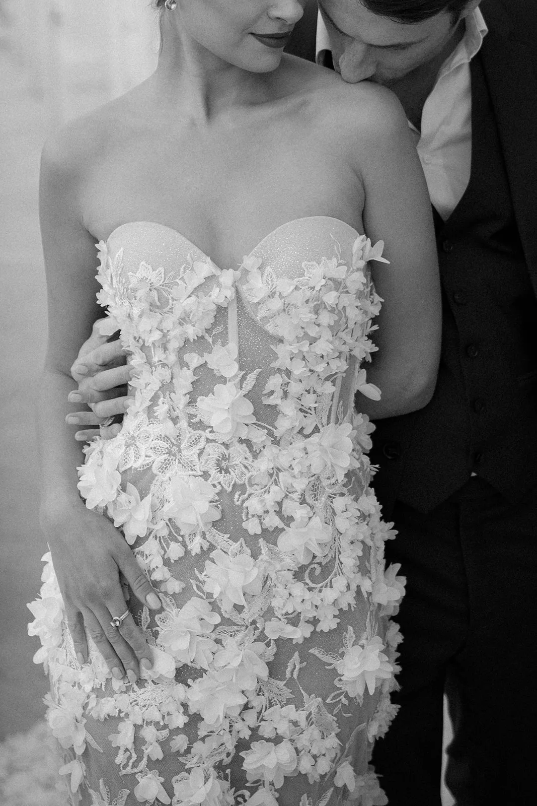 Groom kissing the bride's shoulder above her floral applique gown, in black and white