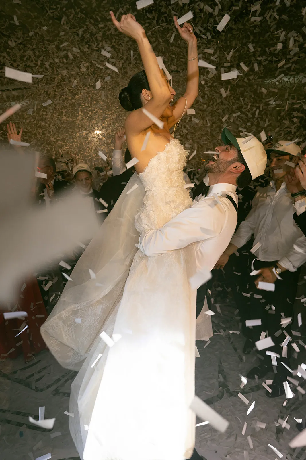 Groom lifting the bride under falling confetti as guests crowd around