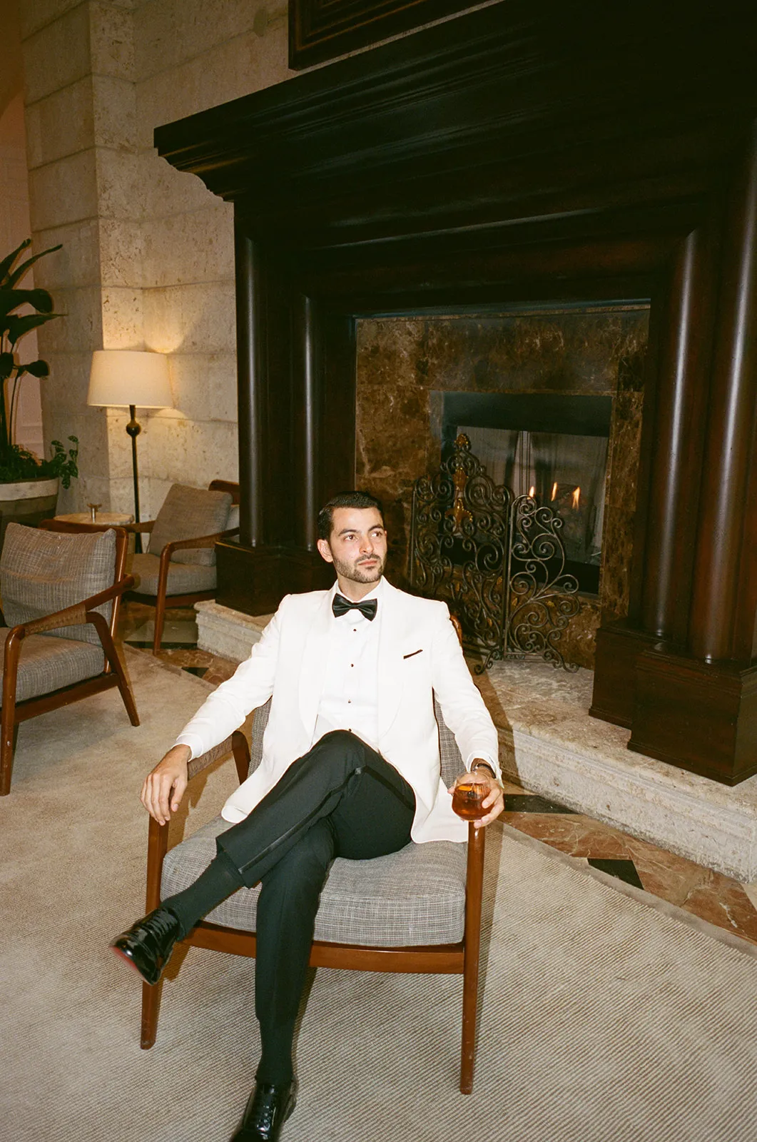 Groom in a white dinner jacket with a cocktail, seated by a lit fireplace