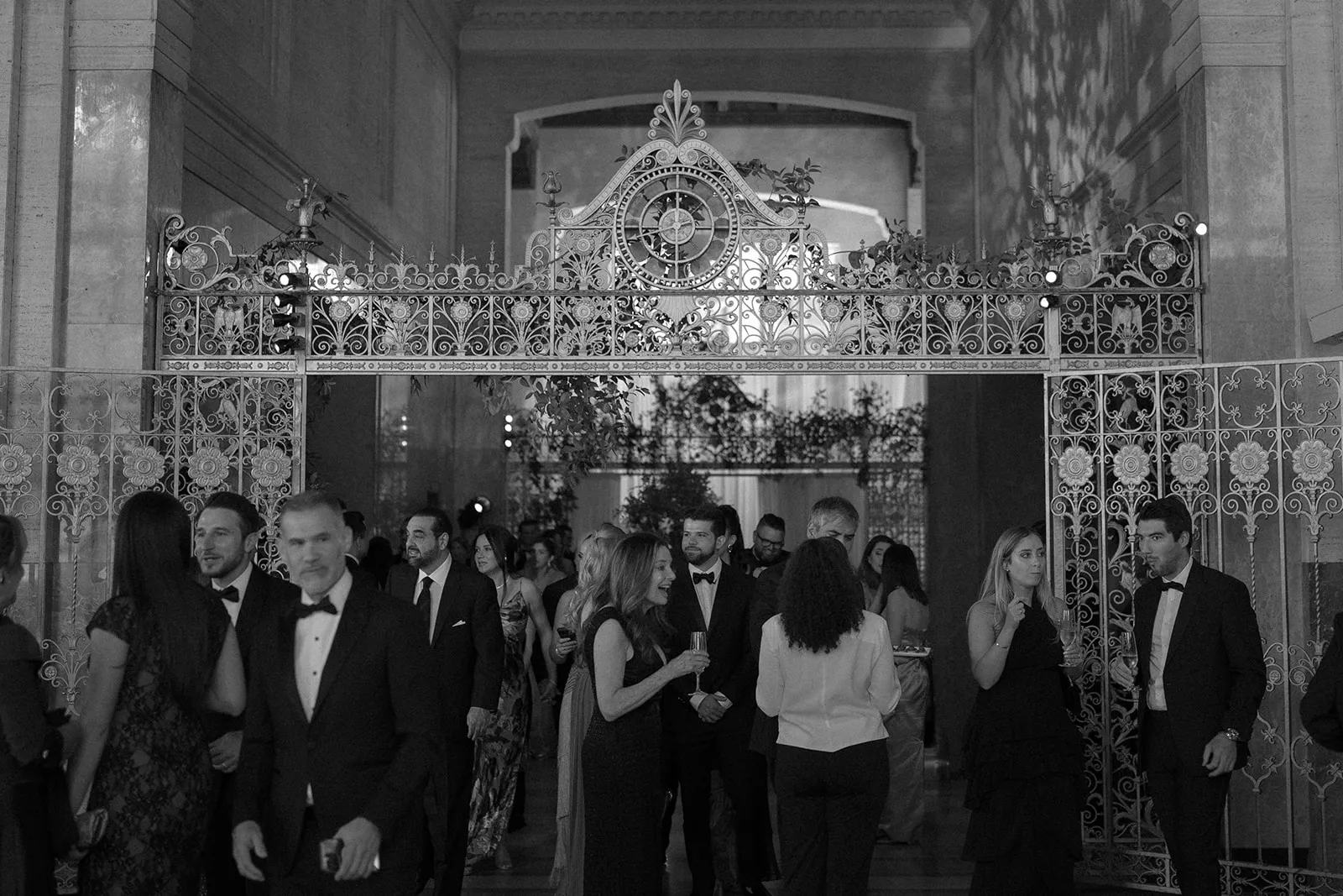 Guests in black tie mingling before an ornate iron gate, in black and white