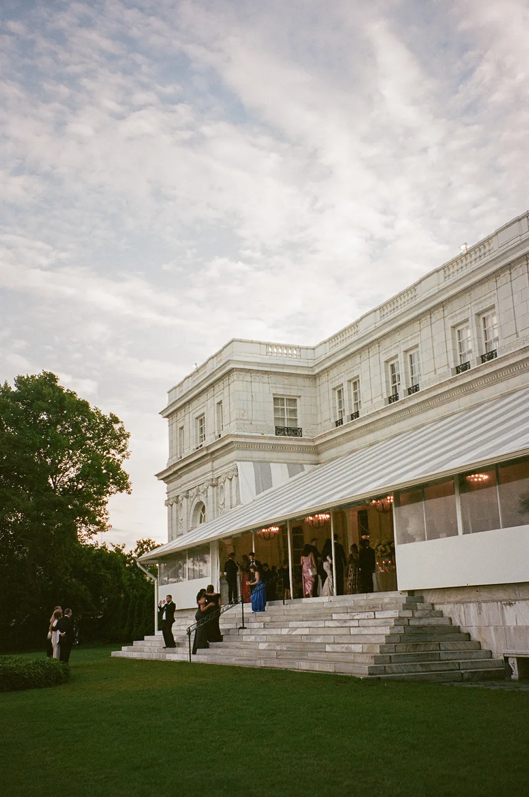 Guests gathering on the marble steps of a white mansion at dusk