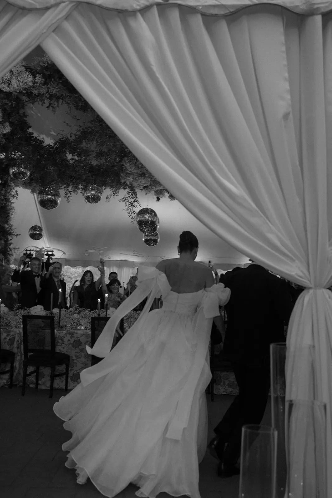 Bride twirling with her veil flying through the curtains of a tented reception, in black and white
