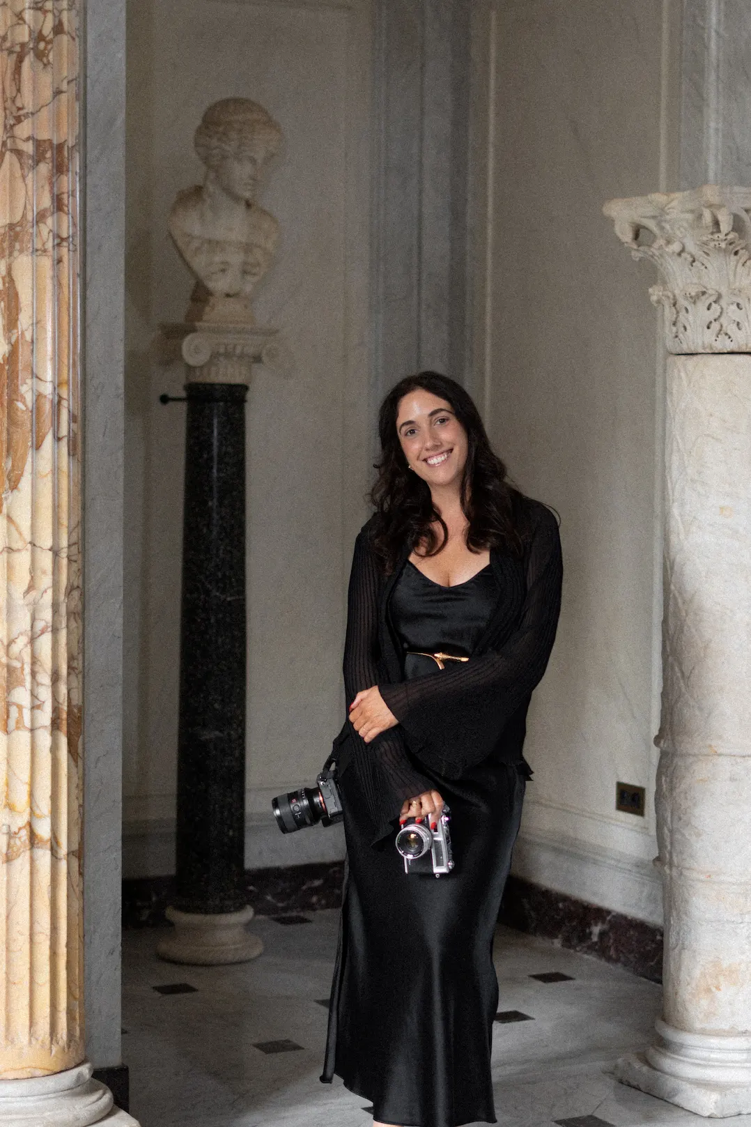 Nadia in a black satin dress, smiling, holding two cameras between marble columns